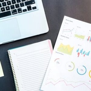 Flatlay of business report with colorful charts, a notebook, and a laptop for data analysis on a desk.